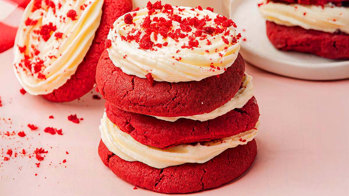 Three stacked red velvet cookies with cream cheese frosting and red crumbs on top, placed on a light pink surface.