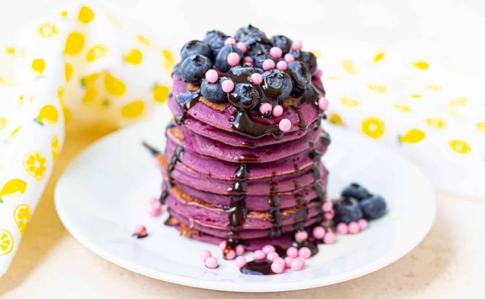 A stack of purple pancakes topped with blueberries, pink candy pearls, and drizzled chocolate sauce on a white plate.