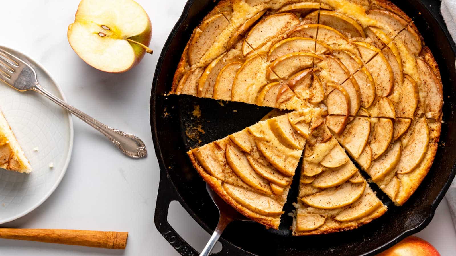 A sliced apple tart in a cast iron skillet, with a serving spatula, fork, and a plate with a slice of tart next to a halved apple on a white surface.