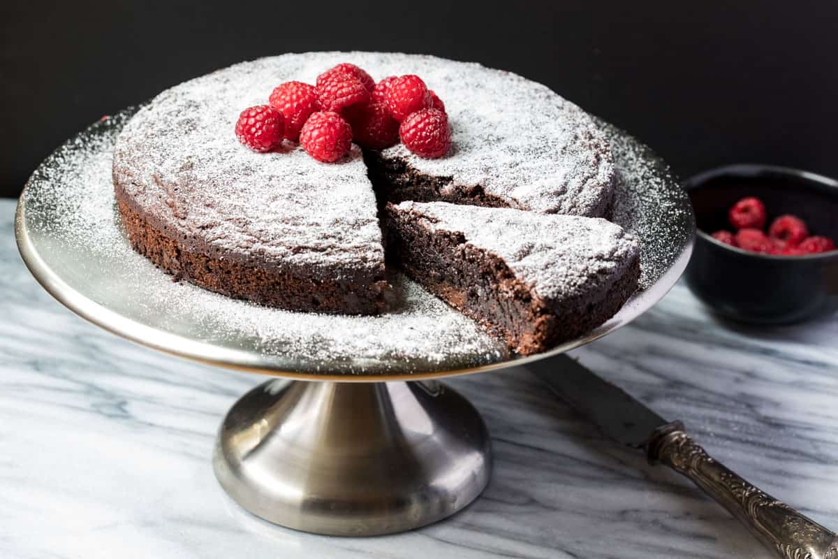 A round chocolate cake dusted with powdered sugar, topped with fresh raspberries, sits on a metal cake stand; one slice is cut and slightly separated.