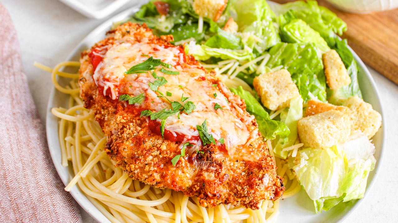 A plate with breaded chicken topped with tomato sauce and cheese on spaghetti, served with Caesar salad and croutons.