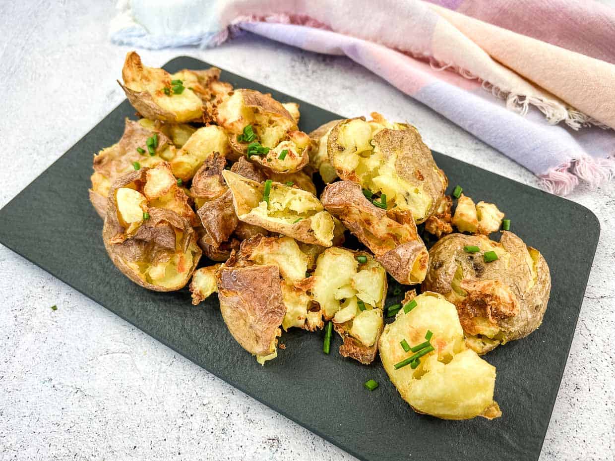 Crispy smashed potatoes topped with chopped chives are served on a black slate platter with a pastel cloth in the background.