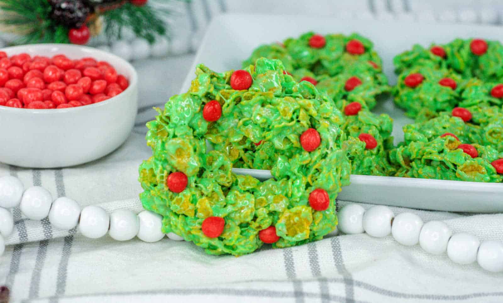 Green cornflake wreath cookies with red candy decorations are displayed on a white plate, with a bowl of red candies in the background.