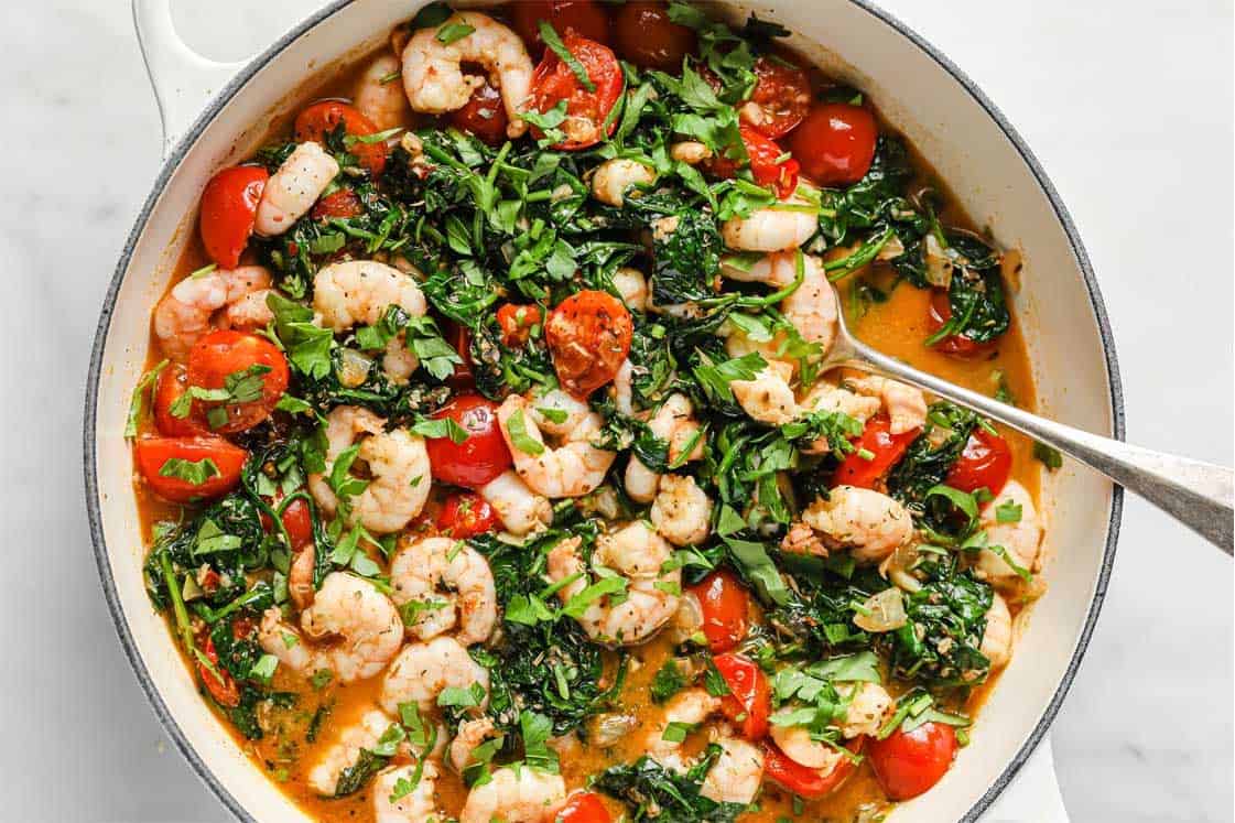 Shrimp cooked with cherry tomatoes, chopped greens, herbs, and a light sauce in a white pan, with a spoon on the side.
