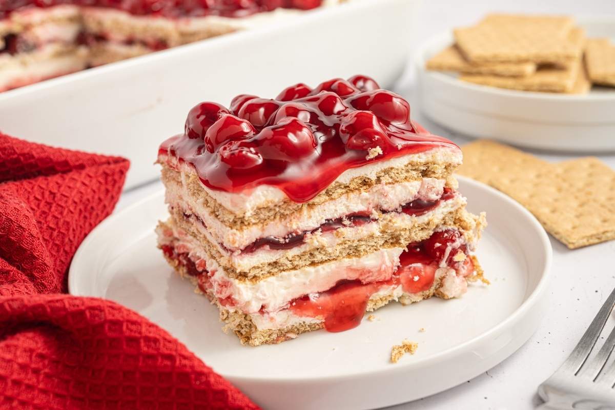 A slice of cherry layered dessert with graham crackers, cream filling, and cherry topping sits on a white plate next to a red cloth and extra graham crackers.