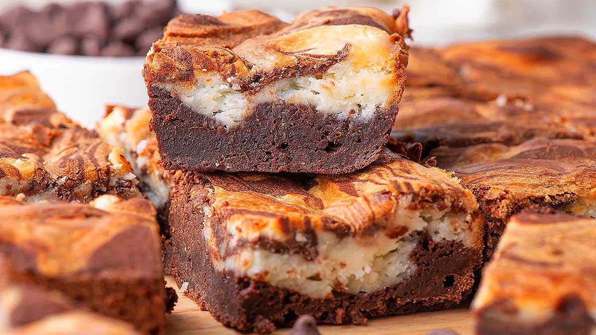 Close-up of marbled brownies with a cream cheese swirl filling, stacked on a wooden surface with more brownies and chocolate chips in the background.