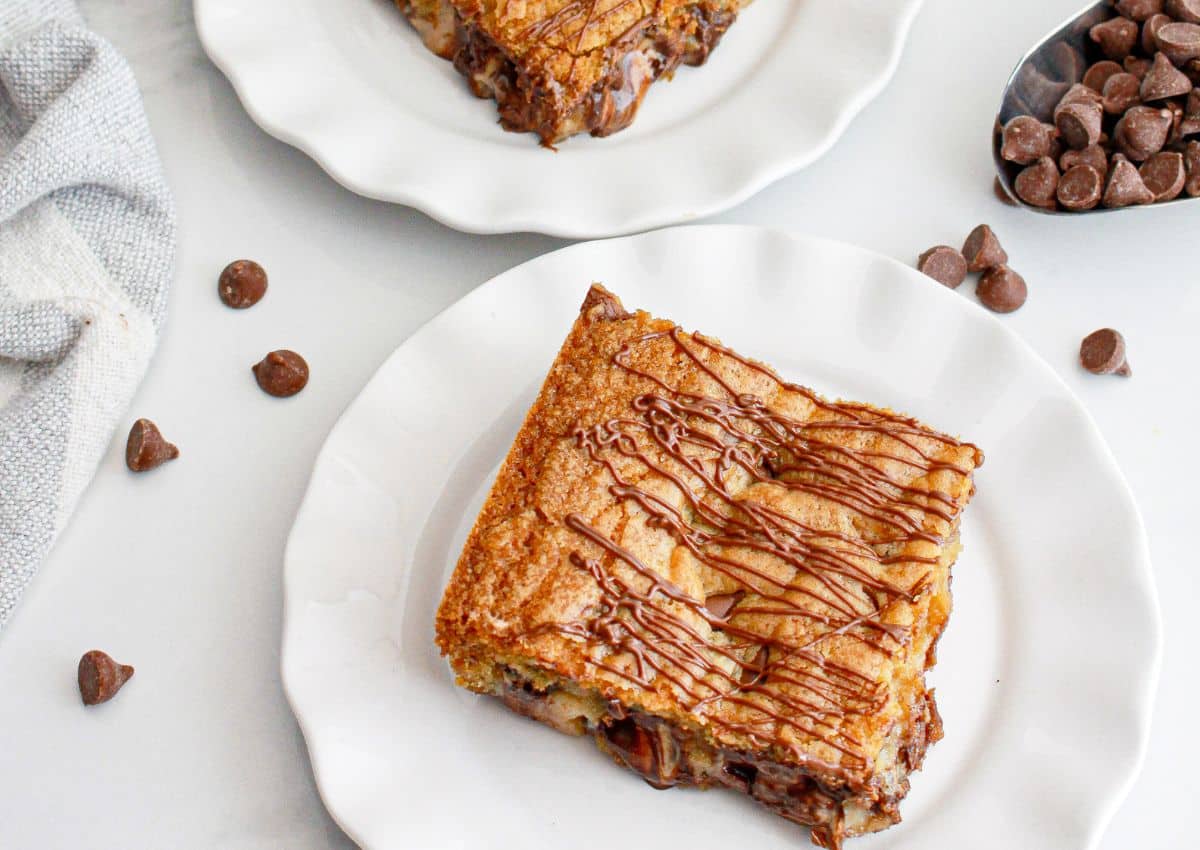 A square chocolate chip blondie drizzled with chocolate on a white plate, with chocolate chips scattered nearby.