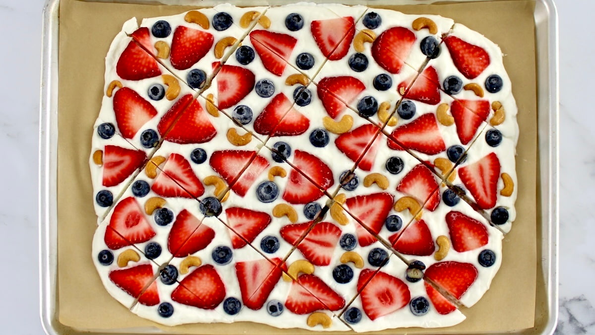Rectangular sheet of fruit pizza topped with sliced strawberries, blueberries, and cashews, cut into square pieces and placed on a baking tray lined with parchment paper.