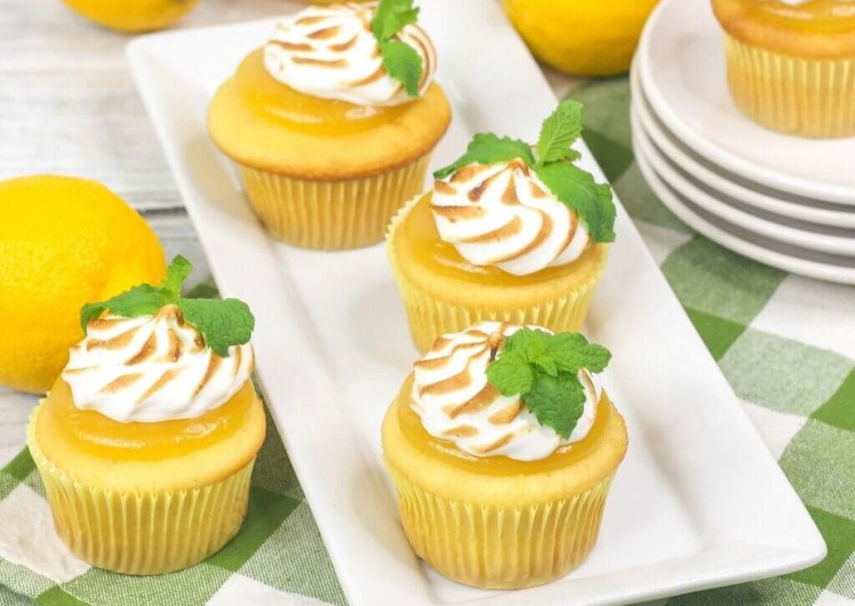 Four lemon cupcakes topped with toasted meringue and mint leaves are arranged on a white rectangular plate, with lemons and stacked plates in the background.