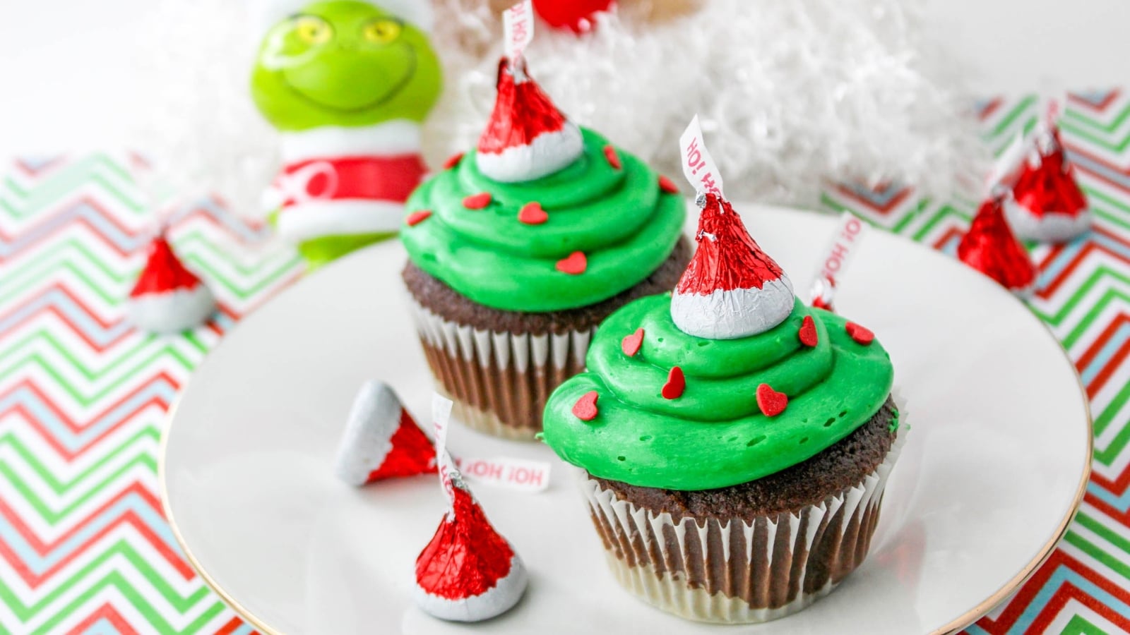 Two chocolate cupcakes with green frosting, red heart sprinkles, and Hersheys Kisses on top, placed on a plate with a Grinch figurine and more Kisses in the background.