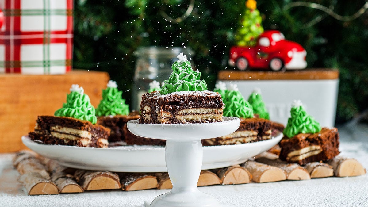 A plate of layered chocolate cakes decorated with green frosting shaped like Christmas trees, placed on a white surface with a festive background.