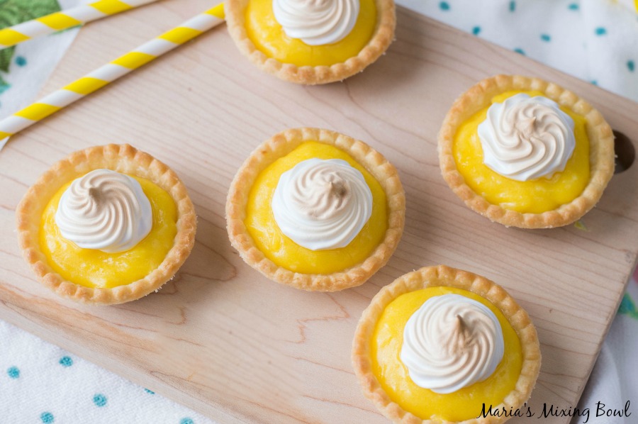 Five mini lemon meringue tarts are arranged on a wooden board, each topped with a swirl of toasted meringue. Two yellow-striped straws are visible in the corner.