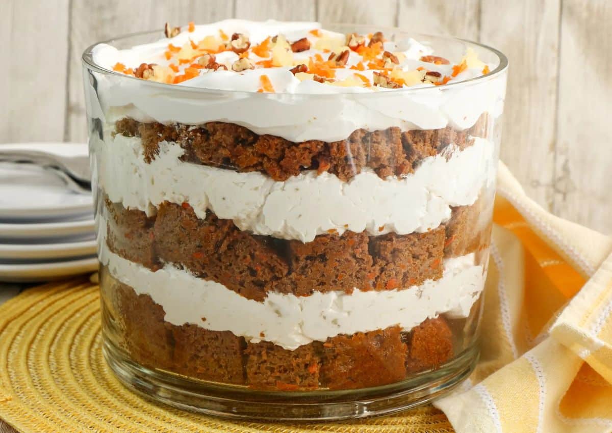 A glass trifle bowl filled with alternating layers of cubed cake, white cream, and topped with chopped nuts, sitting on a woven placemat beside stacked plates.