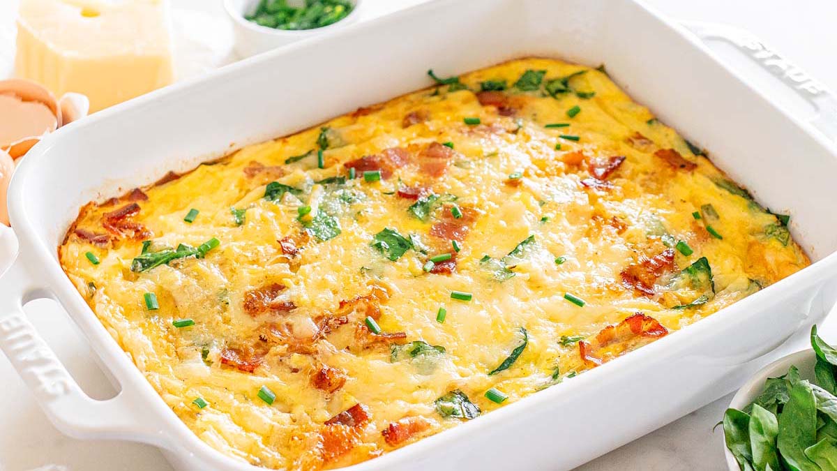 A baked egg casserole with visible pieces of bacon, spinach, and chives in a white rectangular dish.