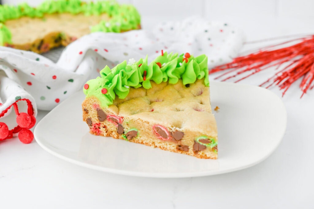 A slice of cookie cake with chocolate and red and green candies, topped with green frosting, on a white plate.