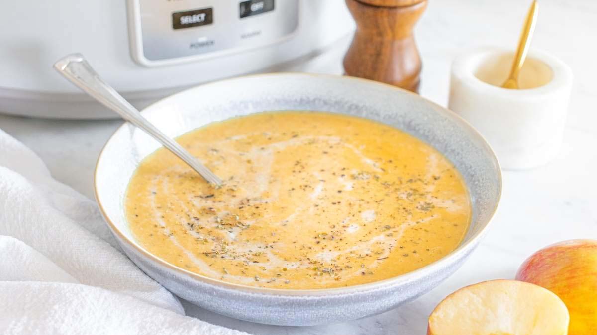 A bowl of creamy orange soup with a swirl of cream and black pepper on top, a spoon in the bowl, and a halved apple, salt, and pepper grinder nearby.