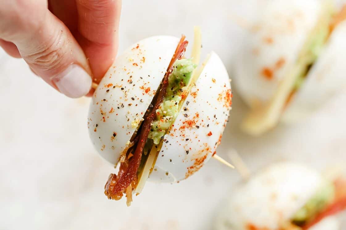 A hand holding a hard-boiled egg sliced in half and filled with avocado, cheese, and bacon, secured with a toothpick and sprinkled with seasoning.