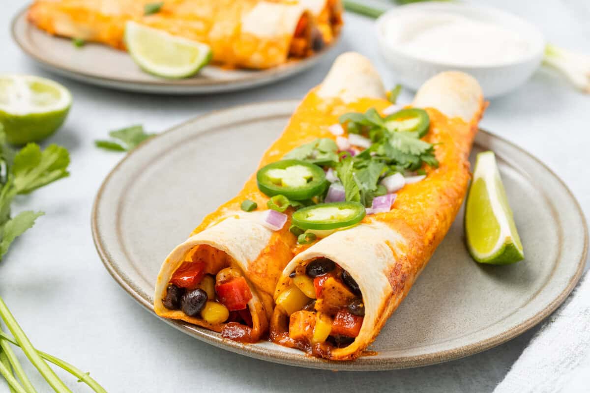 Two vegetable enchiladas topped with jalapeรฑo slices, onions, and cilantro on a plate with lime wedges, with another plate and a bowl of sauce in the background.