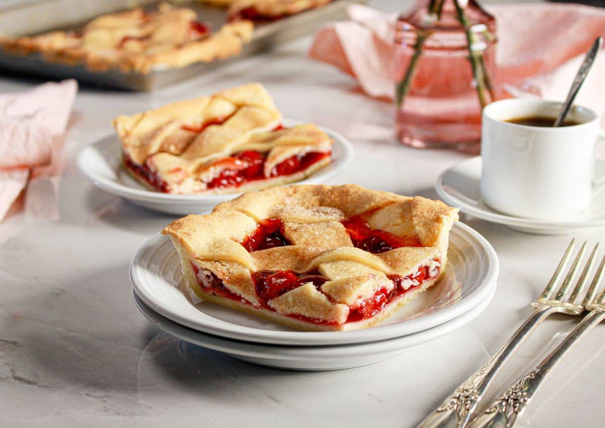 Two plates with slices of cherry pie featuring lattice crust, next to a white coffee cup and silverware on a marble surface.