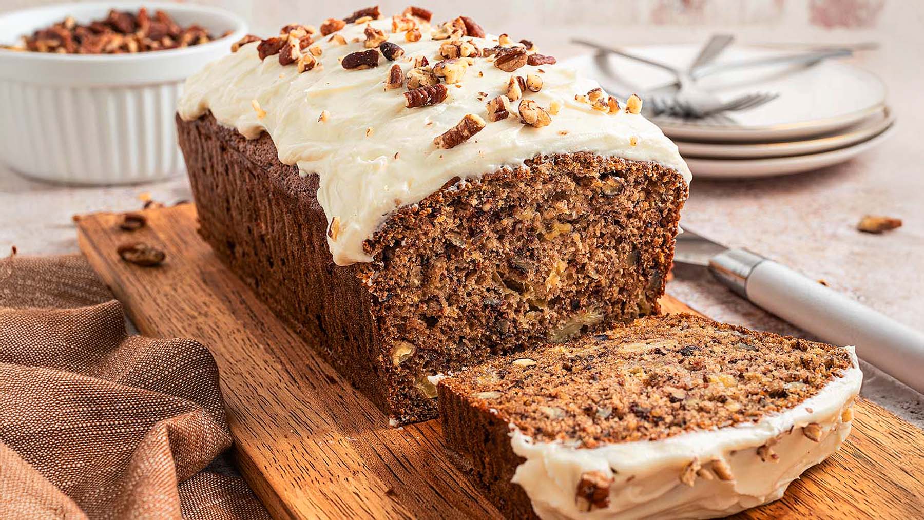 A sliced loaf of banana bread with cream cheese frosting and chopped nuts on top, displayed on a wooden board with a knife and plates in the background.