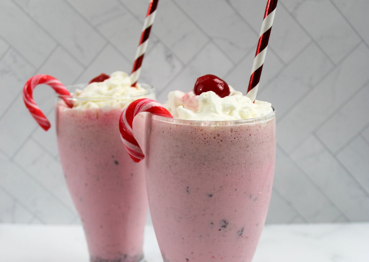 Two pink milkshakes topped with whipped cream and a cherry, each with a candy cane and a striped straw, against a white tiled background.