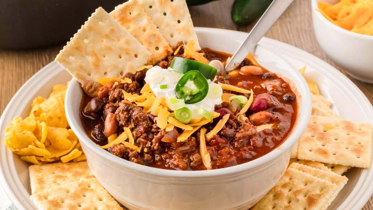 A bowl of chili topped with sour cream, shredded cheese, and sliced jalapeños, served with saltine crackers and tortilla chips on a plate.