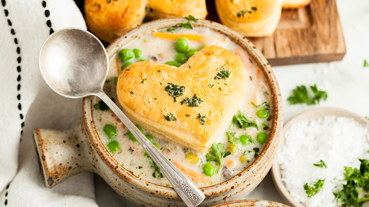 A bowl of creamy soup with peas and carrots, topped with a heart-shaped biscuit; a spoon rests on the bowl and fresh herbs are sprinkled on top.