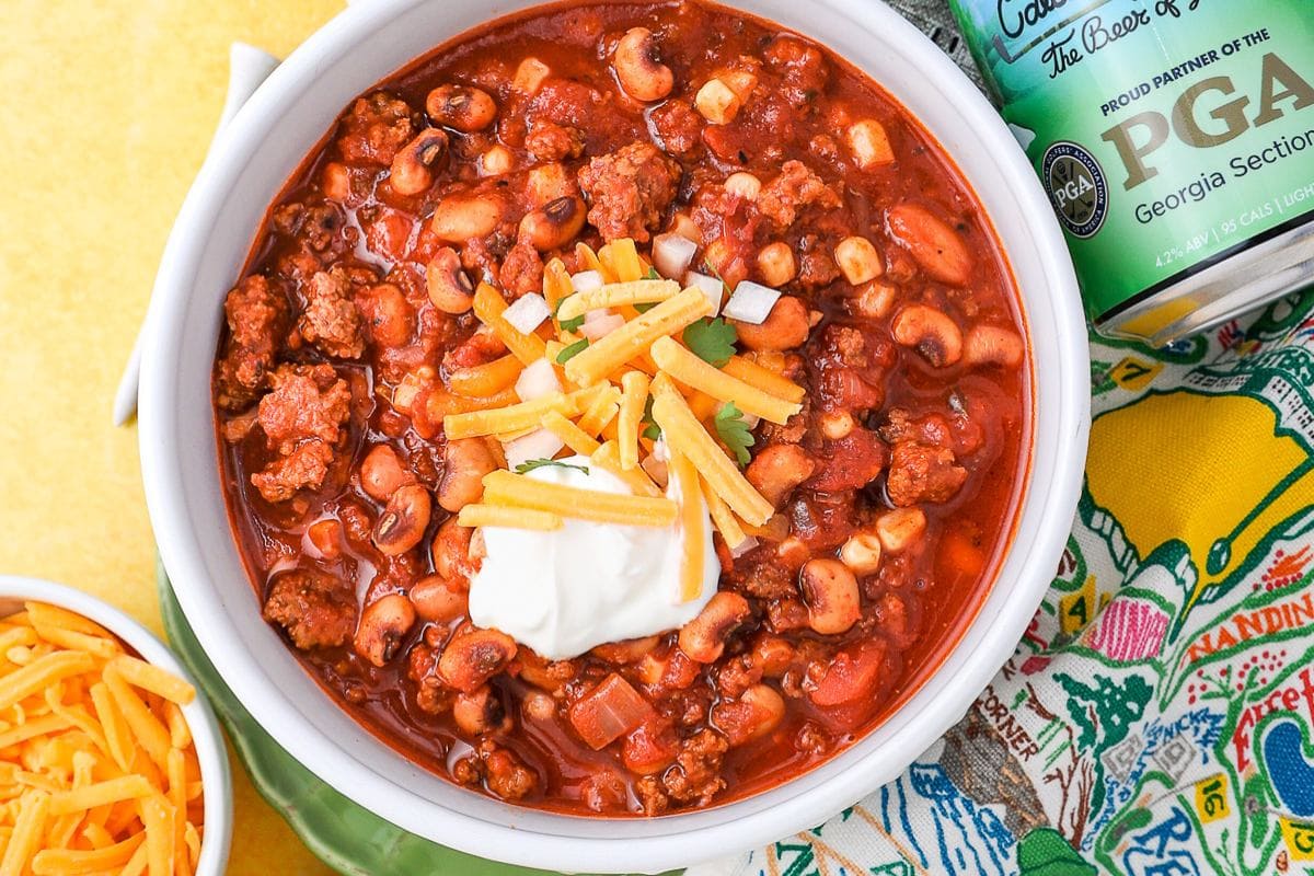 A bowl of chili with ground meat, beans, corn, topped with shredded cheese, sour cream, and chopped onions, next to a can and a bowl of extra cheese.
