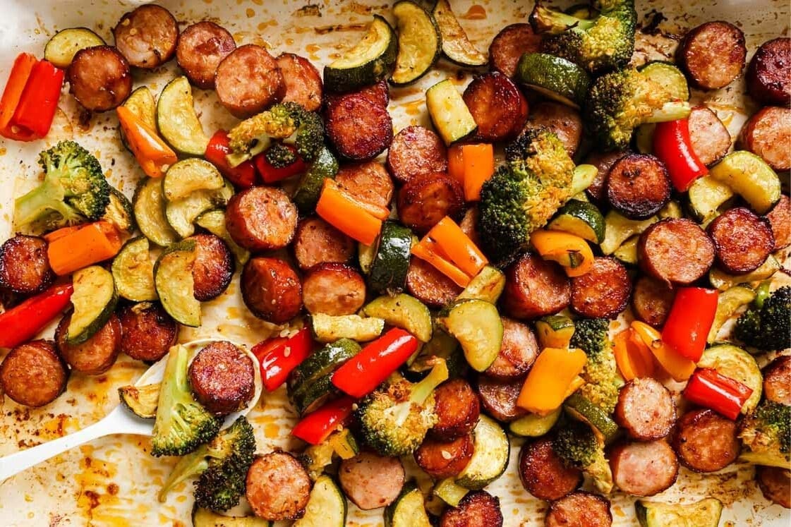 A baked sheet pan meal with sliced sausages, zucchini, broccoli, and red and yellow bell peppers, all roasted and lightly browned.