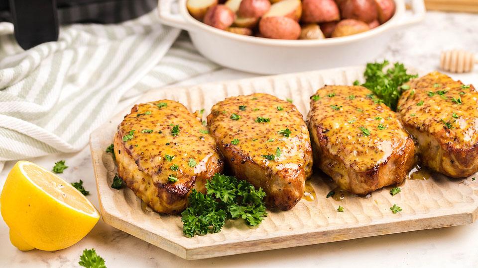 Four glazed pork chops garnished with parsley are arranged on a wooden tray, with lemon halves and a bowl of roasted potatoes in the background.