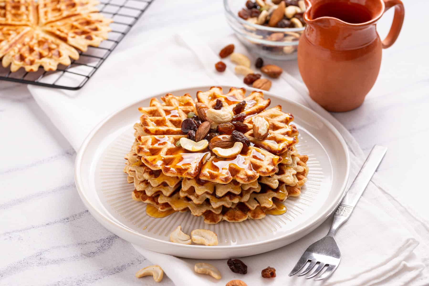 A stack of waffles topped with mixed nuts and raisins sits on a white plate with syrup, next to a fork, napkin, jug, and bowl of nuts.