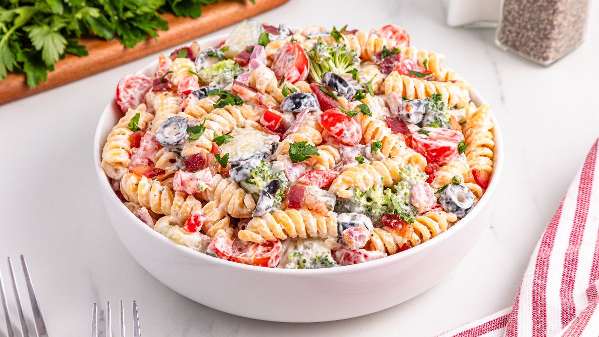 A bowl of rotini pasta salad with cherry tomatoes, broccoli, sliced black olives, red onion, and creamy dressing, garnished with parsley.
