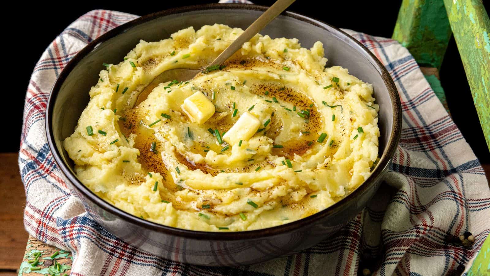 A bowl of mashed potatoes topped with melted butter, chopped chives, and black pepper, with a spoon resting inside, placed on a plaid cloth.