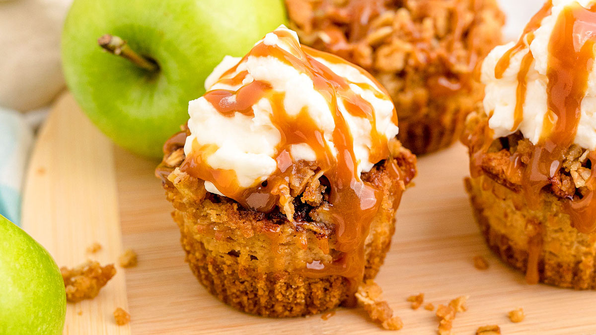 Close-up of apple crumble cupcakes topped with whipped cream and caramel sauce, placed on a wooden board with green apples in the background.