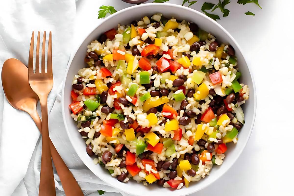 A white bowl filled with a colorful salad of rice, black beans, and chopped red, yellow, and green bell peppers on a white surface with copper utensils beside it.