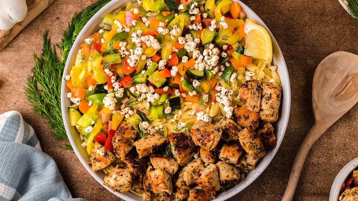A bowl of orzo salad topped with chopped grilled chicken, diced colorful vegetables, white cheese crumbles, and a lemon wedge, with a wooden spoon and herbs on the side.