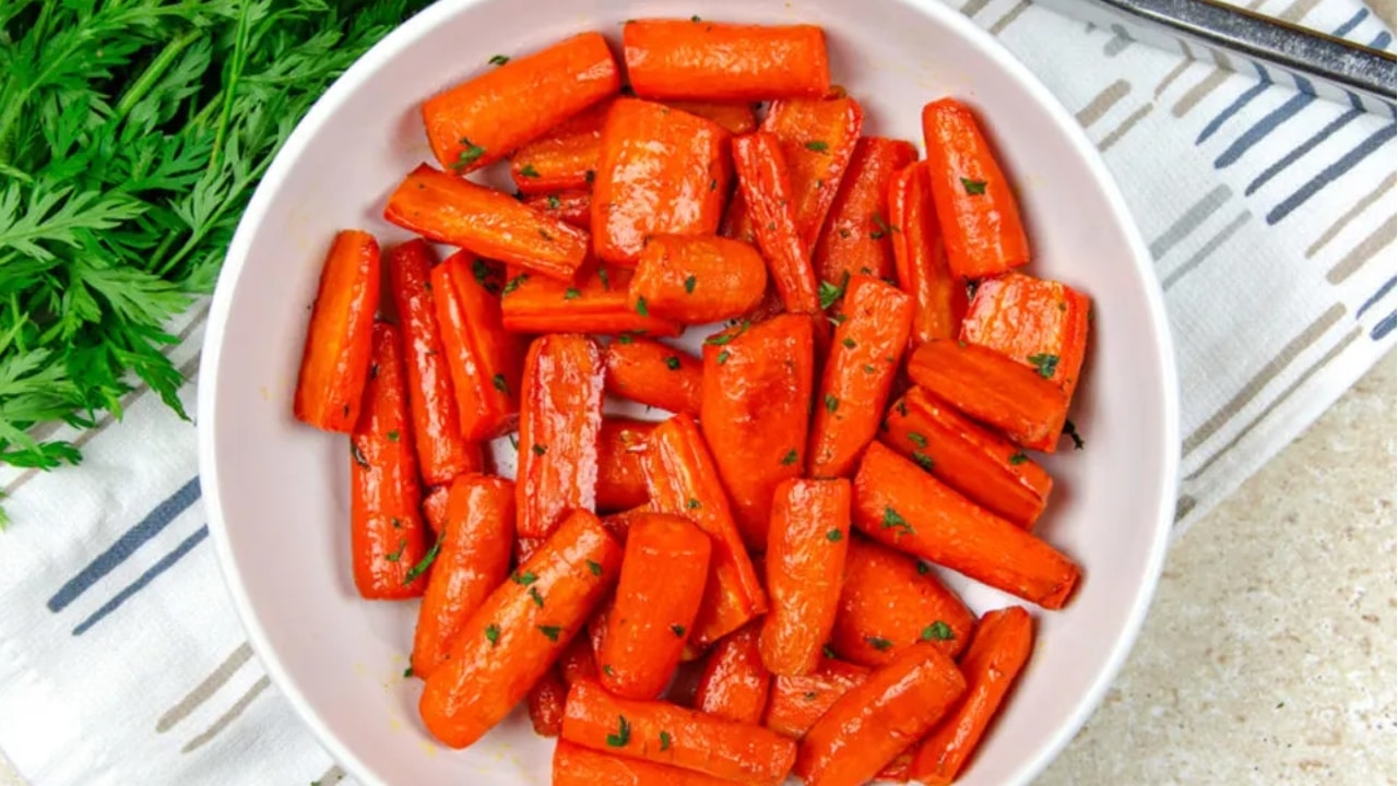 A white bowl filled with roasted carrot pieces garnished with herbs, placed on a striped cloth with carrot greens visible on the side.