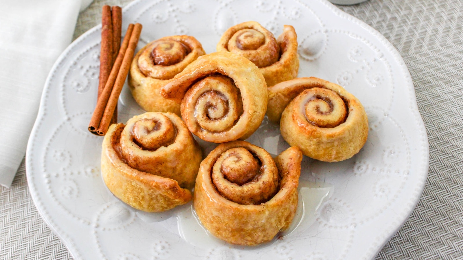 Six cinnamon rolls on a white decorative plate, with two sticks of cinnamon placed beside them.