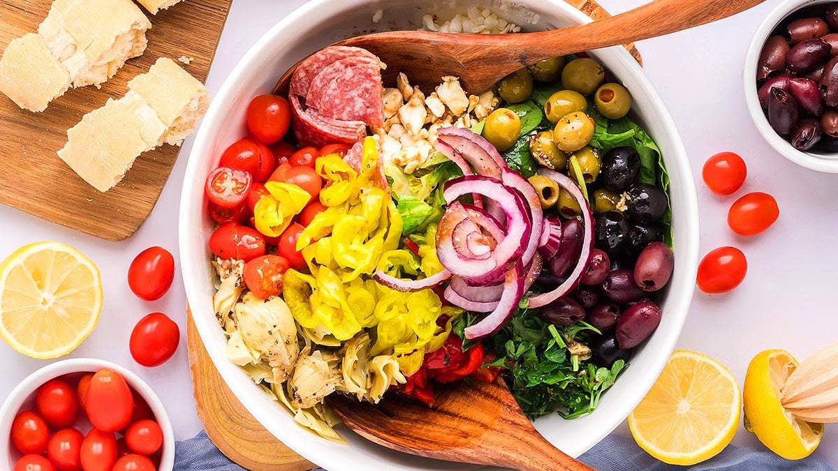 A bowl of salad with cherry tomatoes, olives, salami, sliced red onions, artichokes, pepperoncini, and greens, surrounded by lemon halves, bread slices, and small bowls of olives.