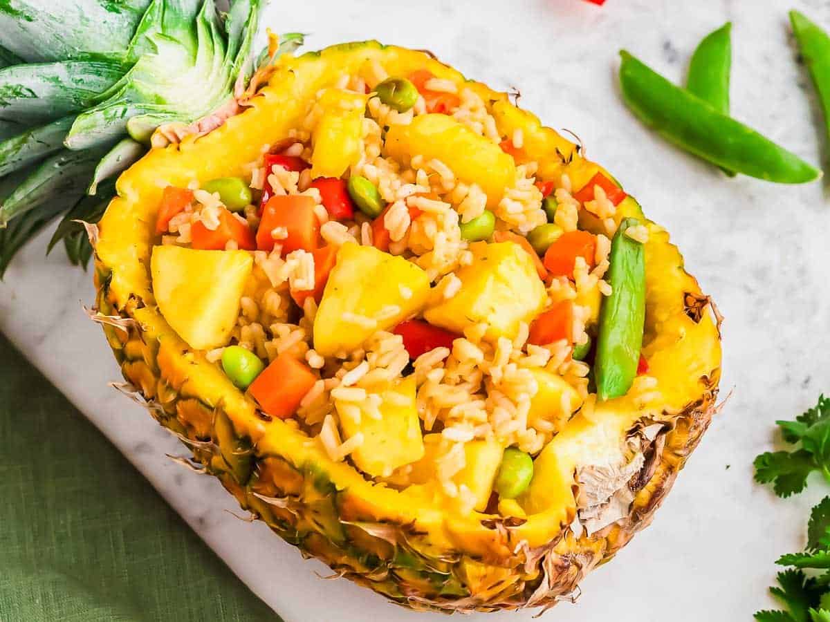 A hollowed-out pineapple half filled with fried rice, pineapple chunks, peas, carrots, and green beans sits on a white surface.