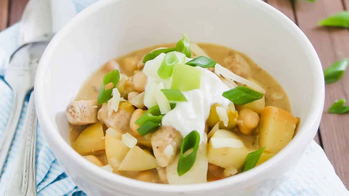 A white bowl filled with a stew containing chickpeas, diced potatoes, and chicken, topped with sour cream, green onions, and shredded cheese.