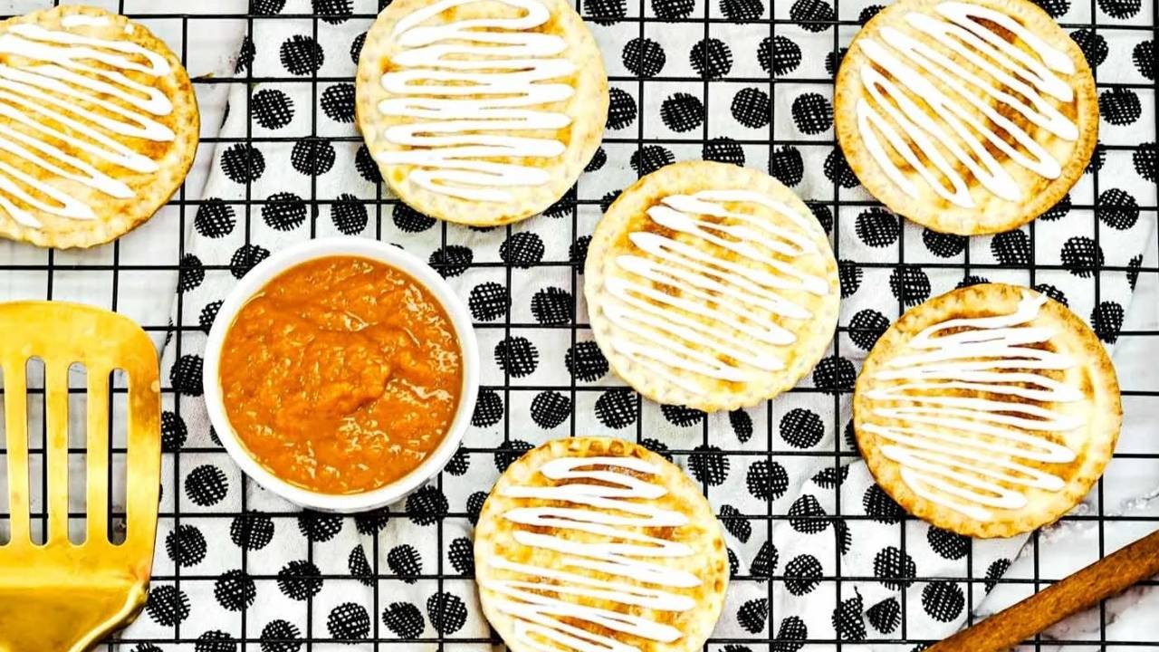 Round pastries with white icing are on a cooling rack, accompanied by a small bowl of orange filling, a gold spatula, and a cinnamon stick.