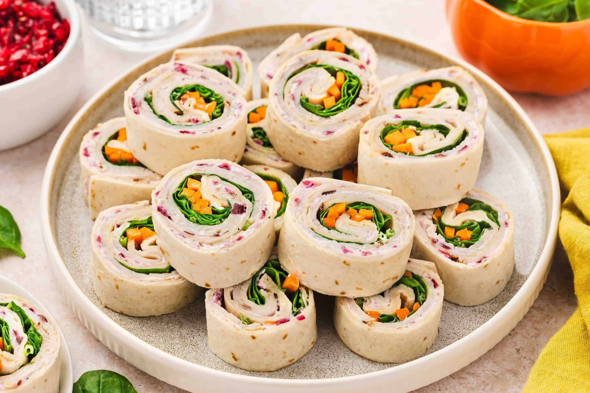 A plate of tortilla pinwheel sandwiches filled with sliced vegetables and leafy greens, arranged neatly in a circular pattern.