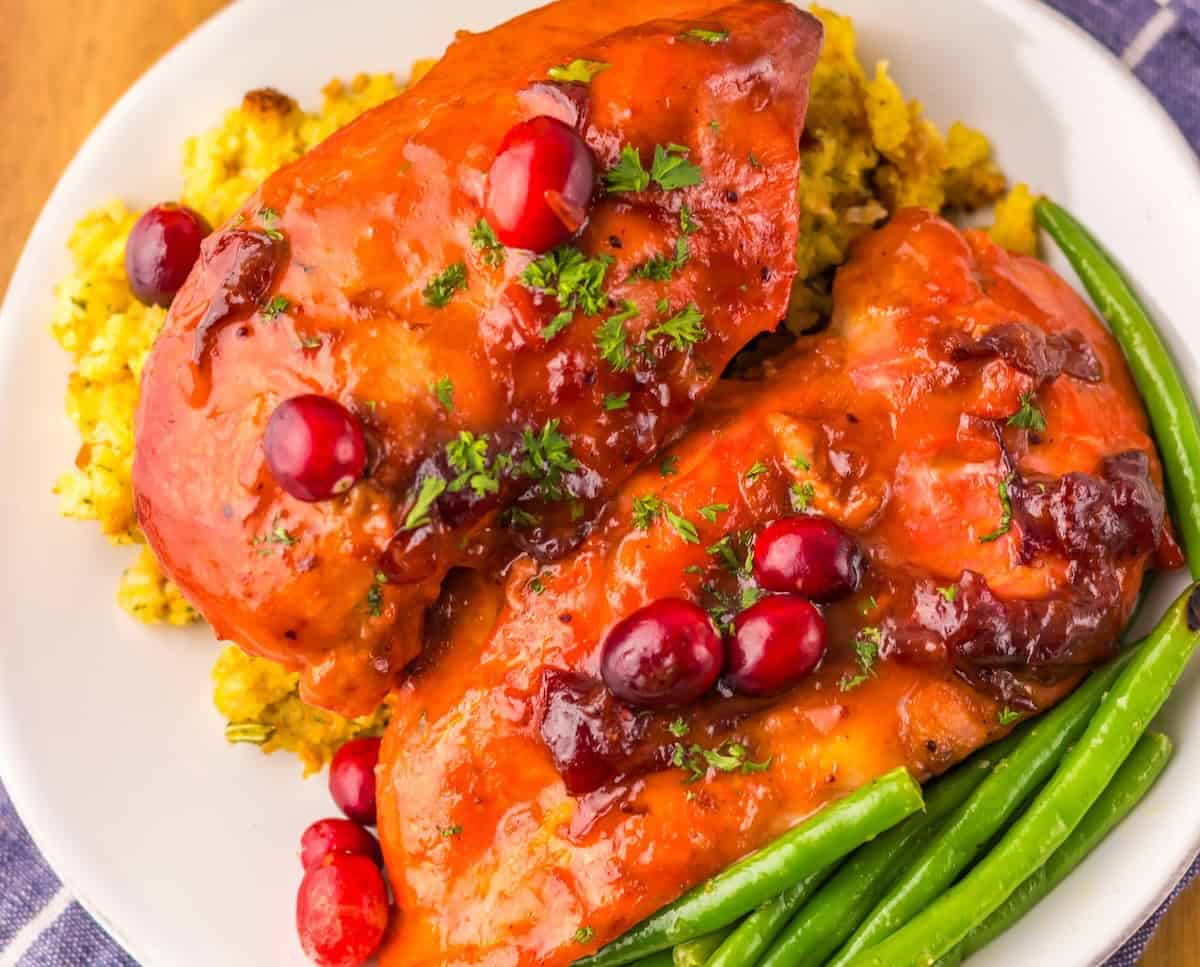 Two glazed chicken breasts topped with cranberries and herbs, served on a bed of rice with green beans on the side.