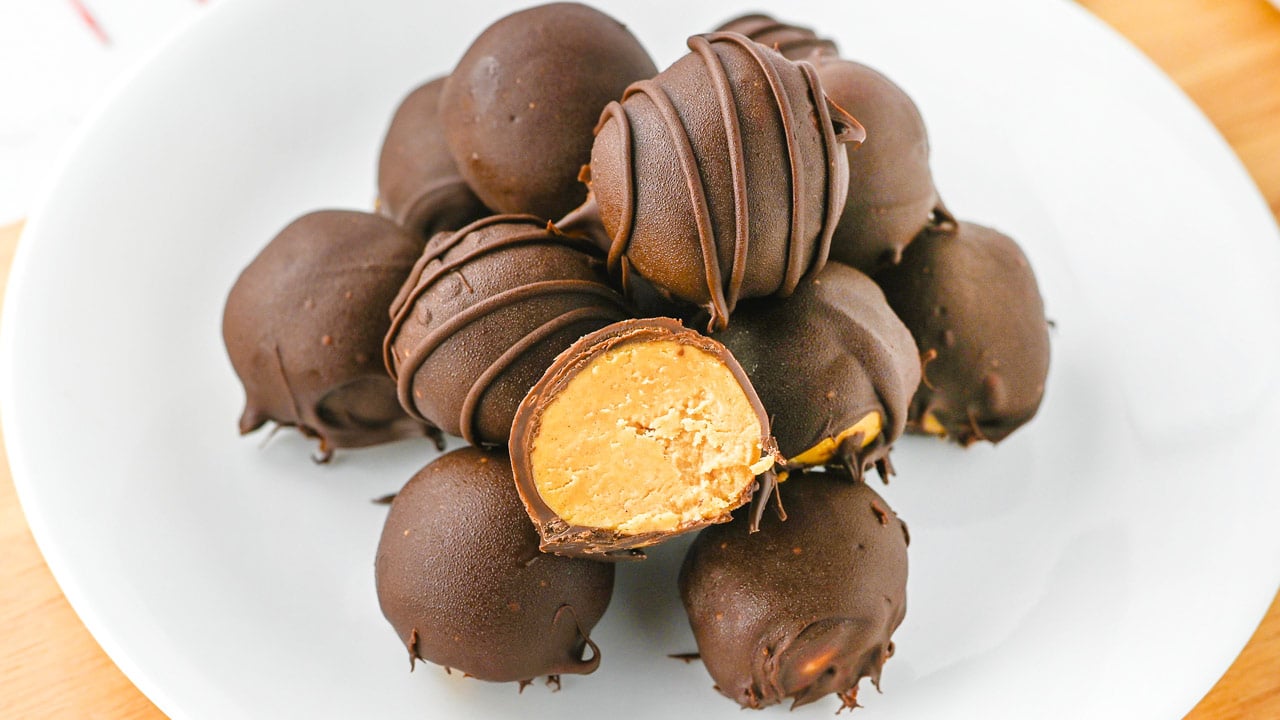 A white plate holds a pile of round chocolate-coated candies, one of which is cut open to reveal a creamy peanut butter filling.