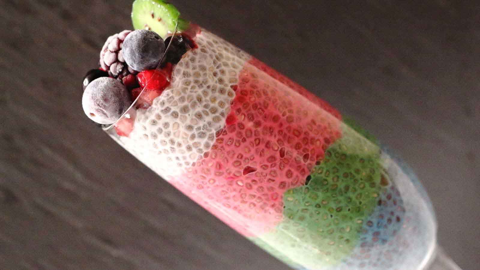 A tall glass filled with layered chia pudding in white, pink, green, and blue, topped with sliced kiwi and assorted frozen berries.