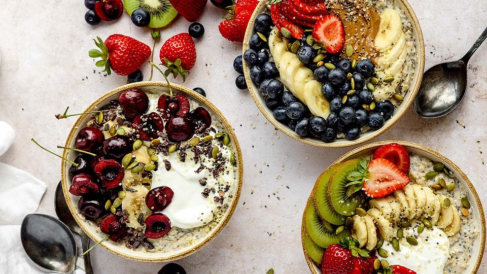 Three bowls of oatmeal topped with fresh fruit, seeds, and yogurt, surrounded by strawberries, kiwi slices, blueberries, and spoons on a light surface.