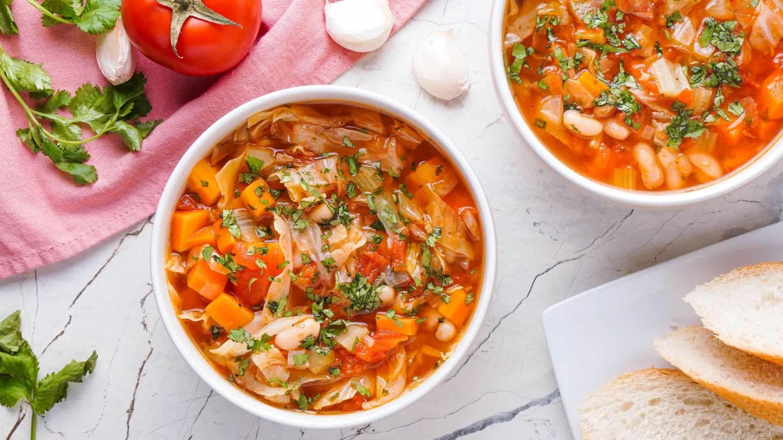 Two bowls of vegetable soup with beans, carrots, and herbs on a marble surface, next to sliced bread, a tomato, garlic, and fresh cilantro.
