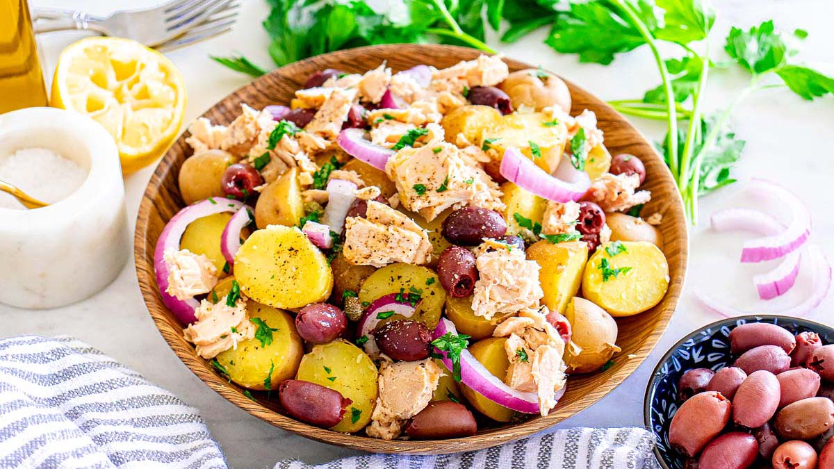 A bowl of potato salad with tuna, kalamata olives, red onion slices, and parsley garnished with a lemon half and fresh herbs in the background.