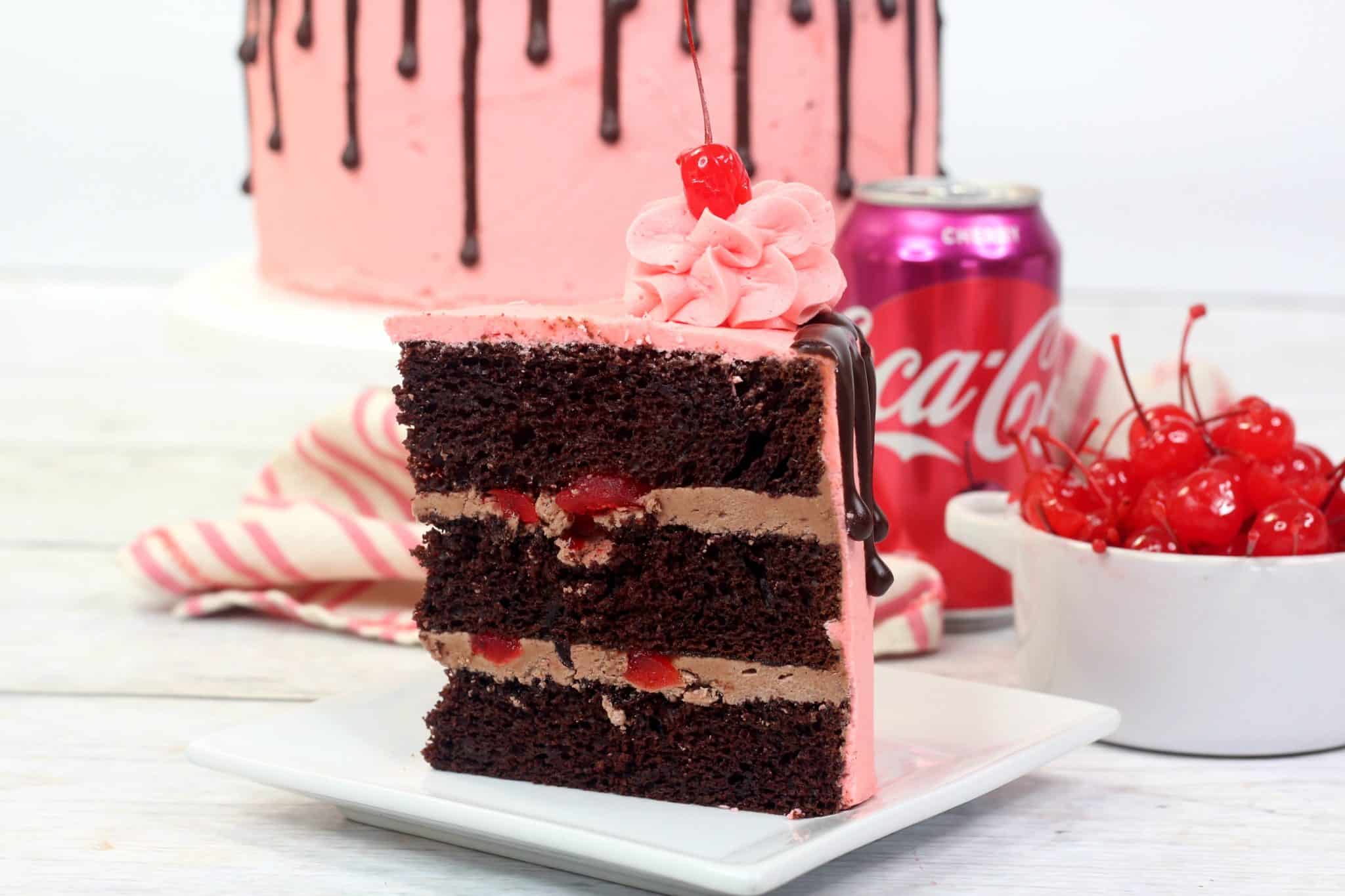 A slice of chocolate cake with pink frosting and a cherry on top sits on a plate, with a can of Cherry Coca-Cola and a bowl of maraschino cherries in the background.
